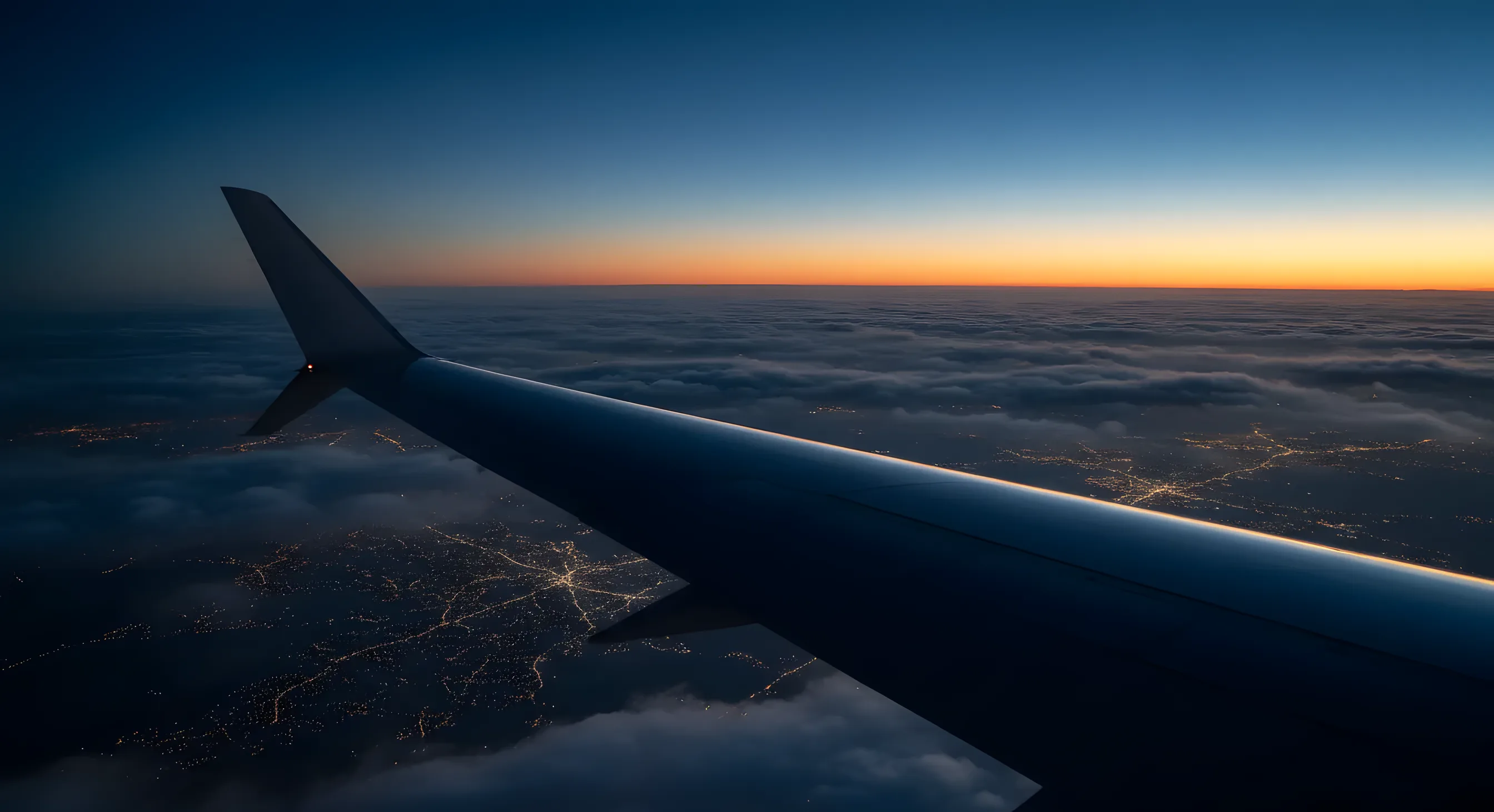 View from an airplane window at sunset, showing the wing over a layer of clouds with city lights visible below.