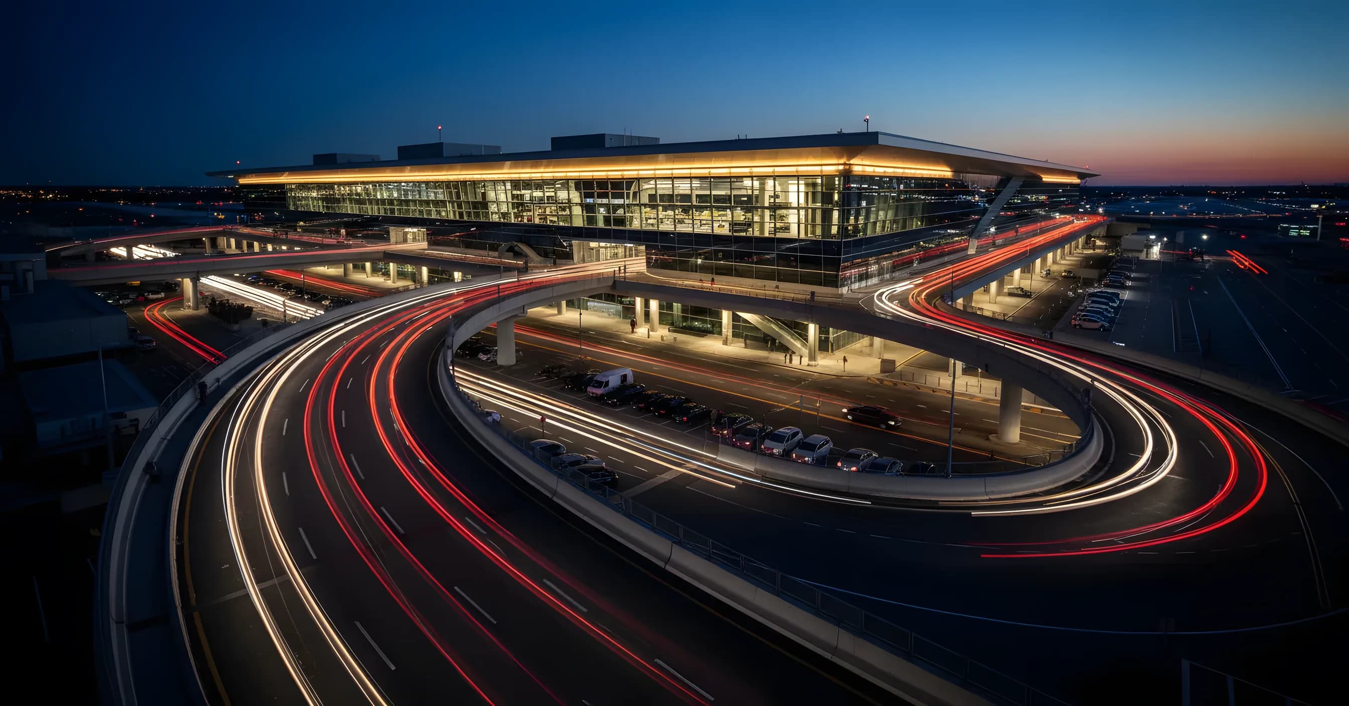 A modern airport terminal glows with warm lights at dusk, with streaks of car lights on the roadway in front.