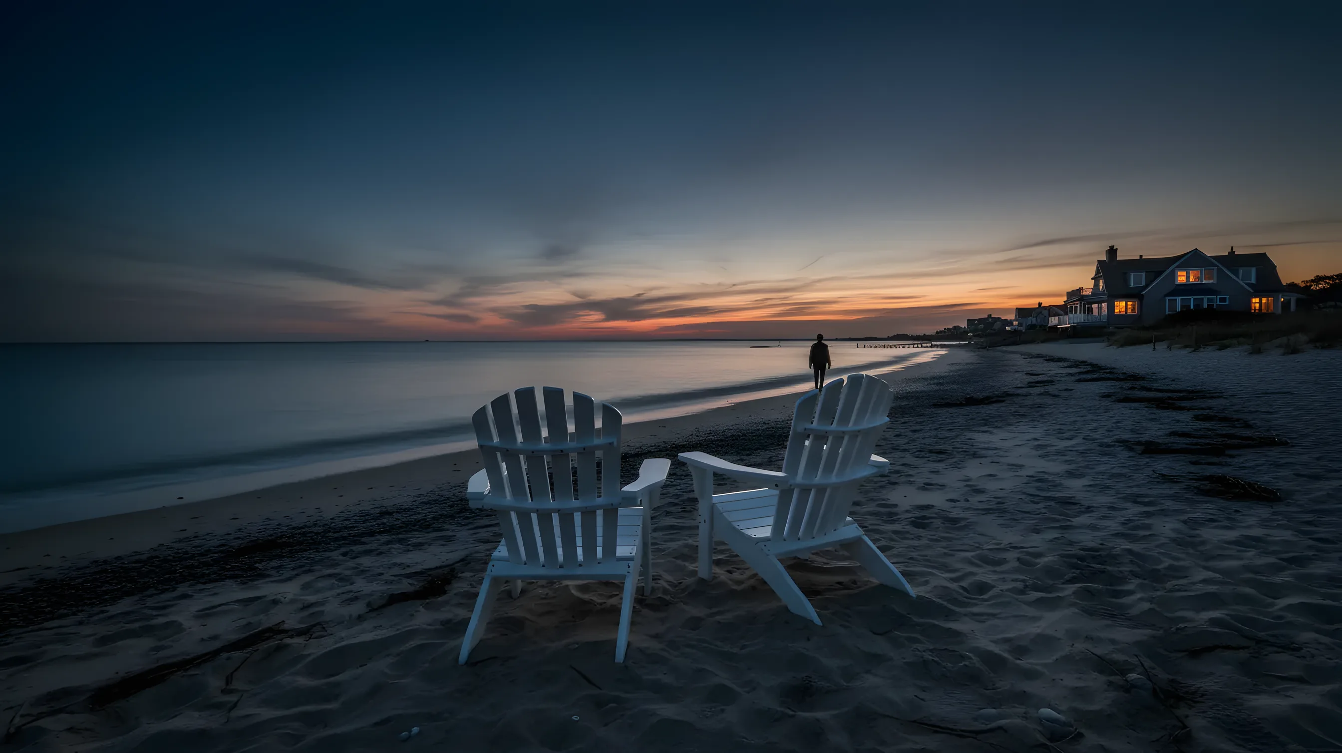 Two empty adirondack chairs on a sandy beach facing the ocean at sunset.