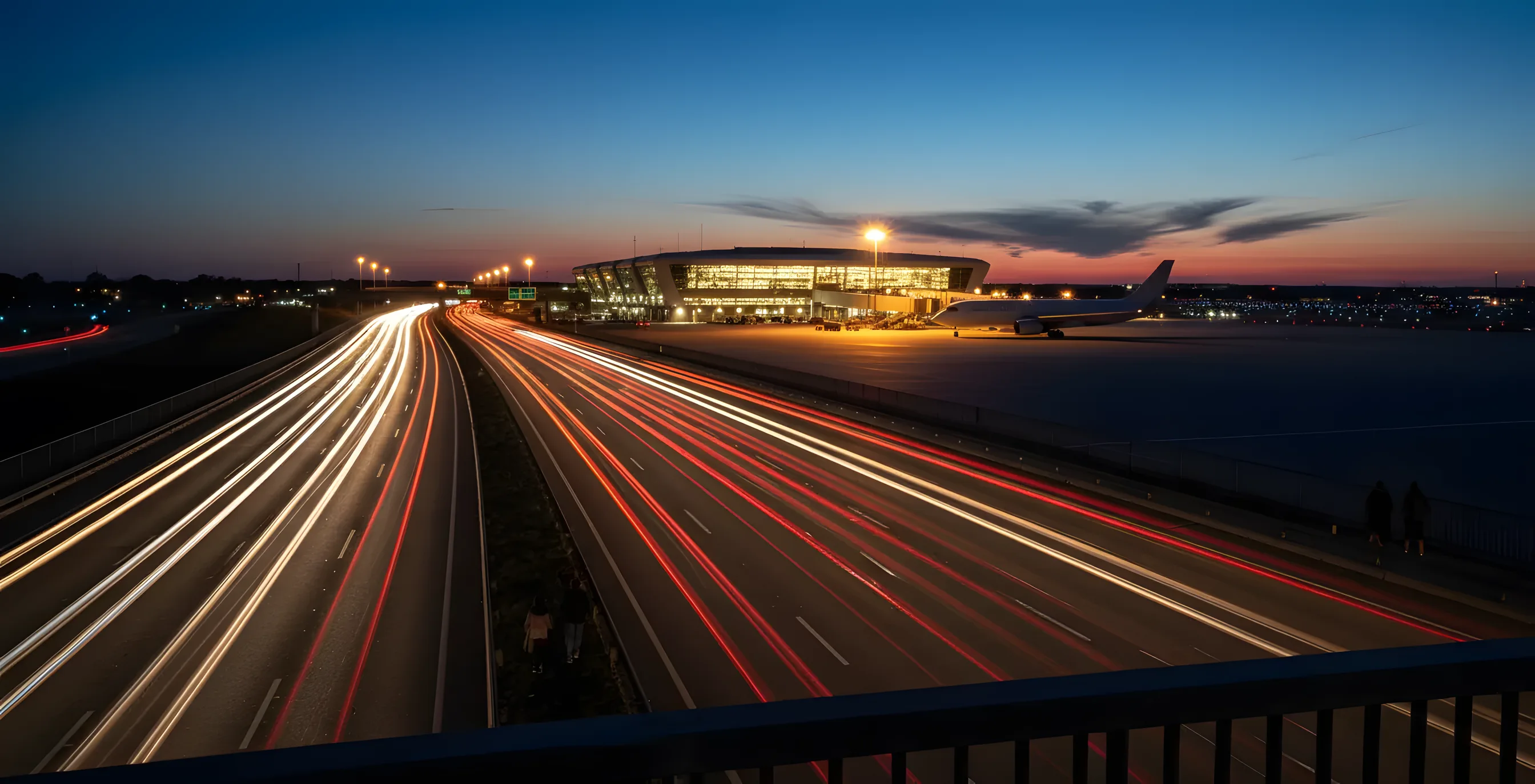 Car light trails on a highway at dusk leading to an airport terminal with a passenger jet on the tarmac.