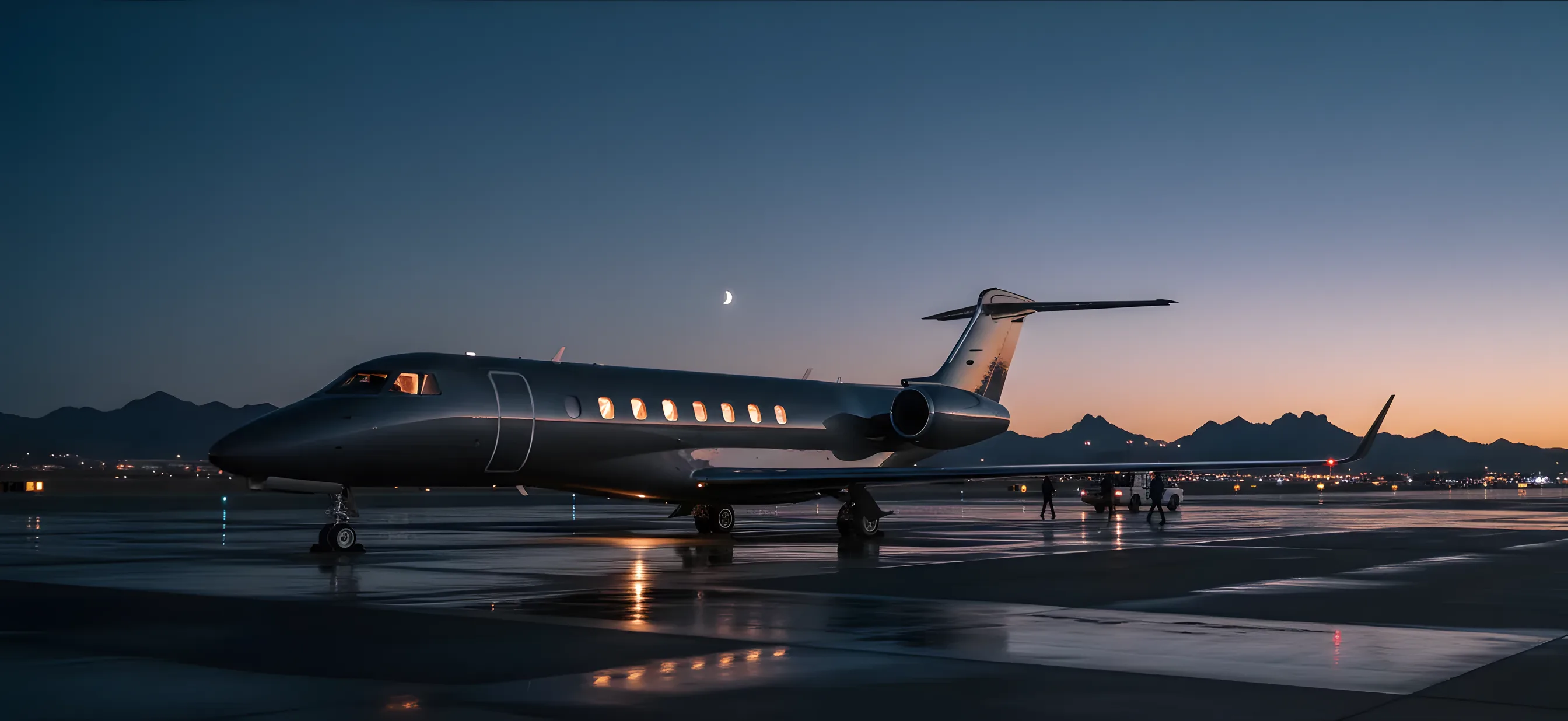 A private jet on an airport tarmac at dusk, with cabin lights glowing against a twilight sky and distant mountains.