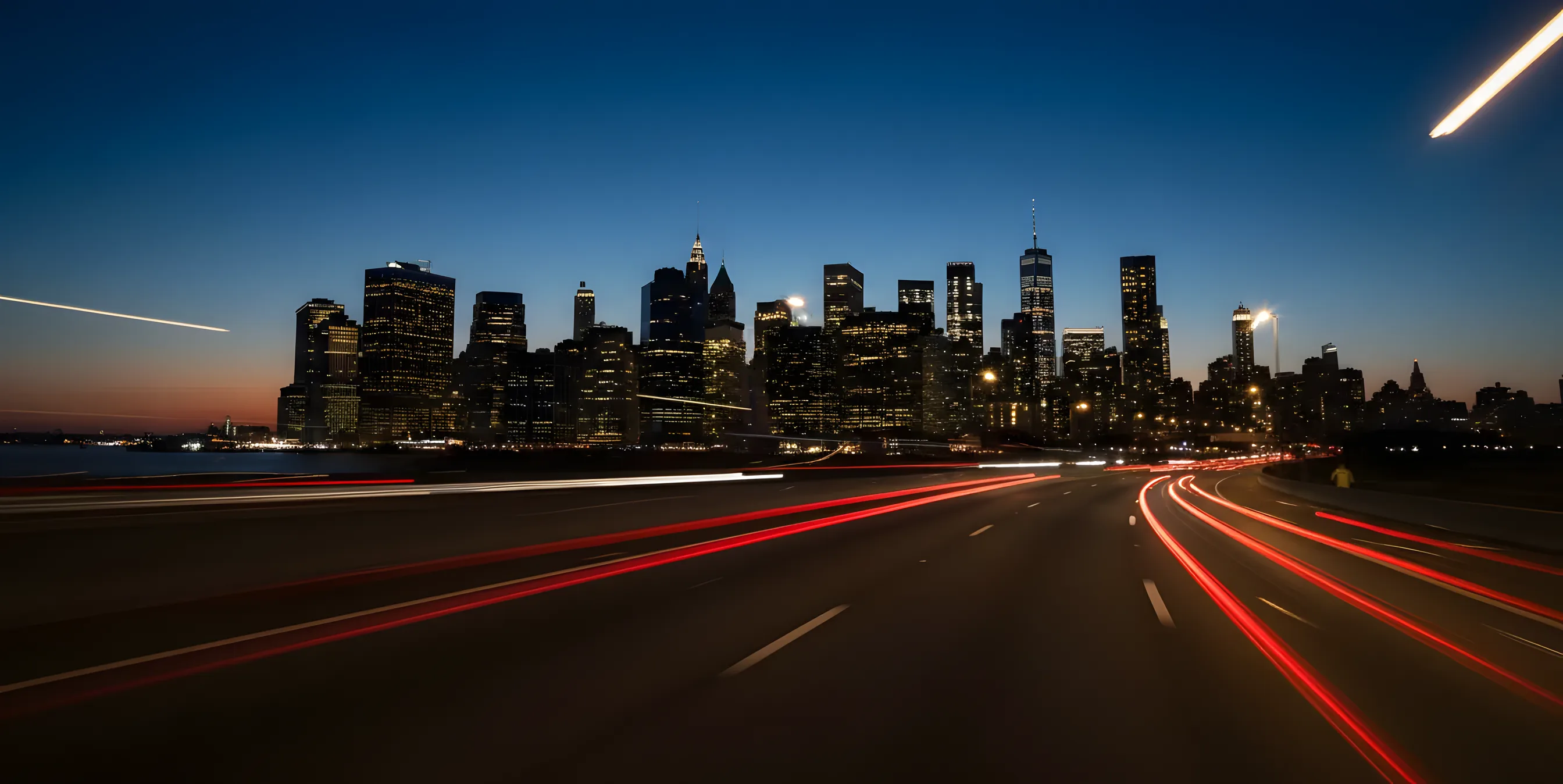 The Manhattan skyline glows with amber lights at night, viewed from a highway with light trails from moving cars.