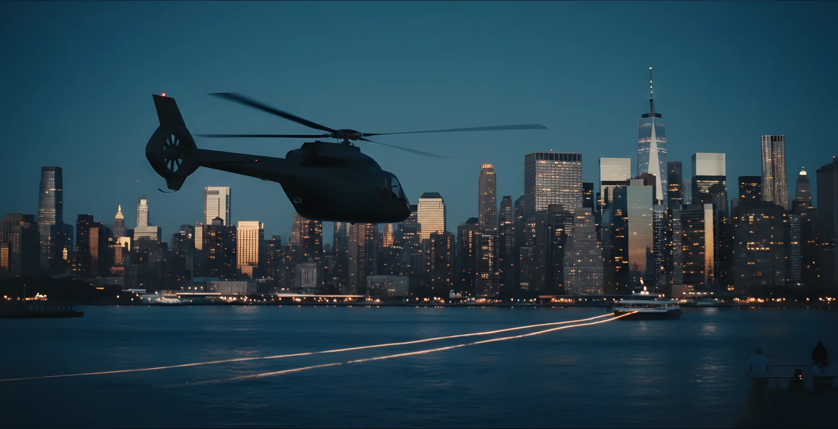 A helicopter flies over a river at dusk, with a brightly lit city skyline in the background.