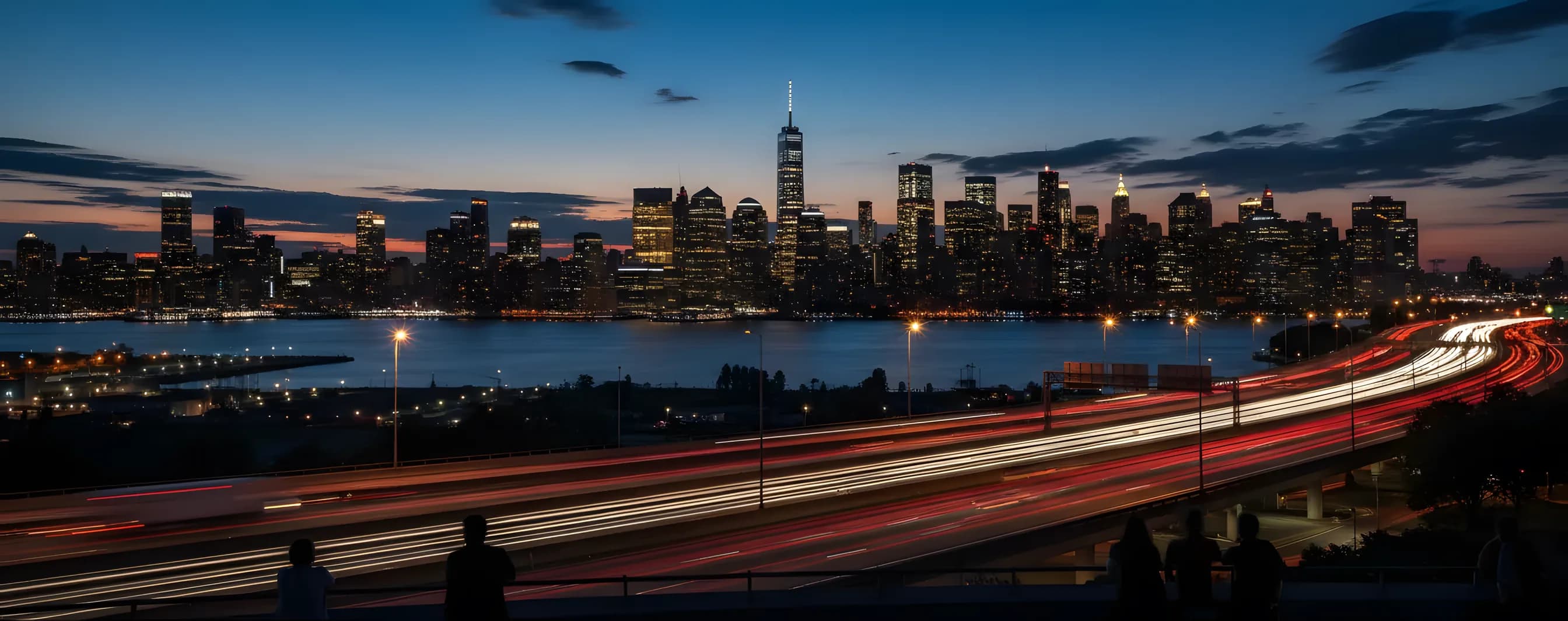 The Manhattan skyline glows with lights at dusk, viewed from across the river with streaks of highway traffic in the foreground.