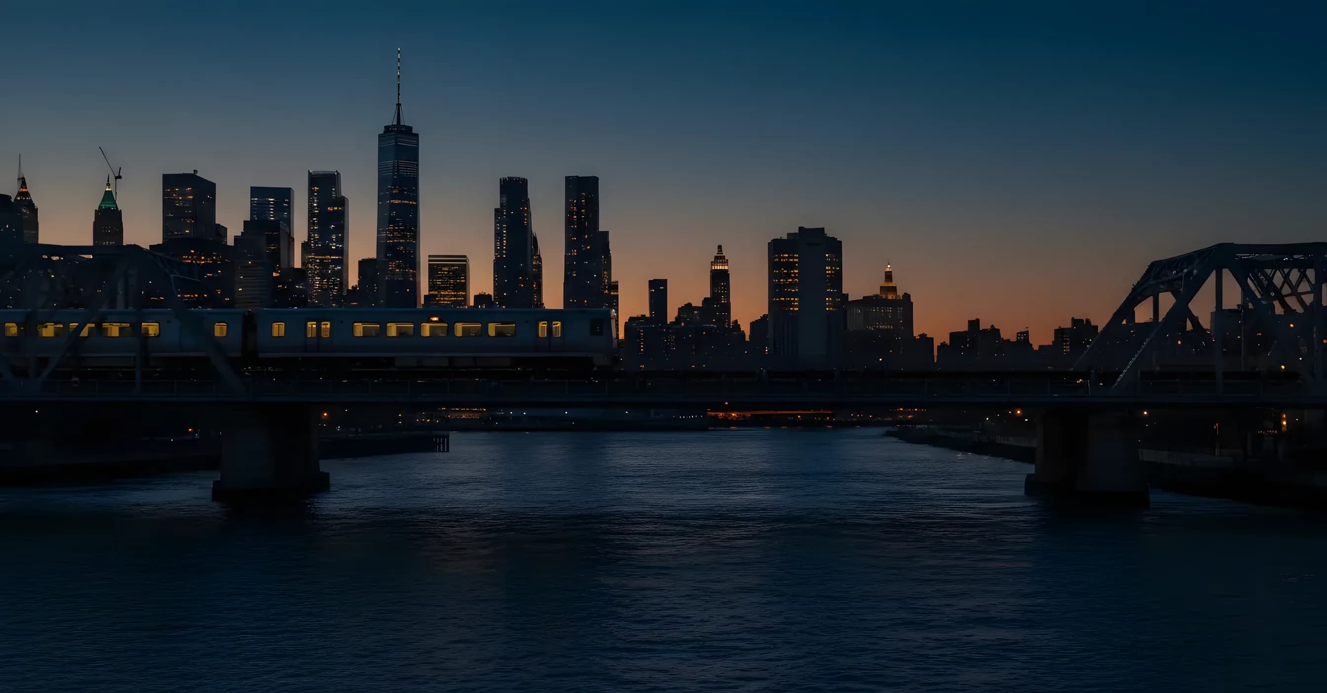 A modern train crosses a bridge at dusk with a glowing city skyline in the distance.