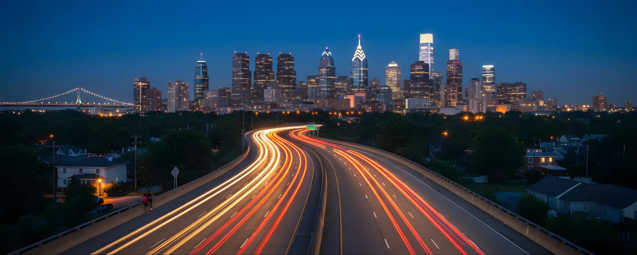 A highway with light trails from traffic at dusk, leading toward the glowing Philadelphia skyline.