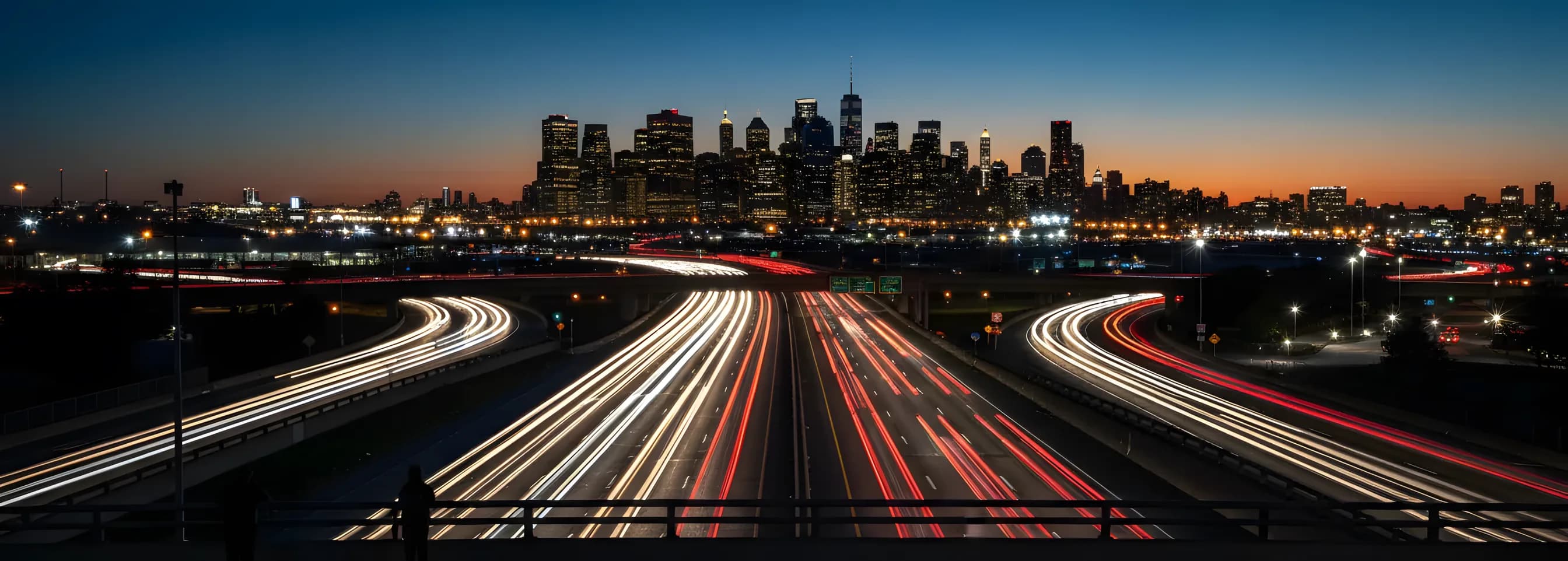 Streams of car lights on a highway leaving an airport at dusk, with a glowing city skyline in the distance.