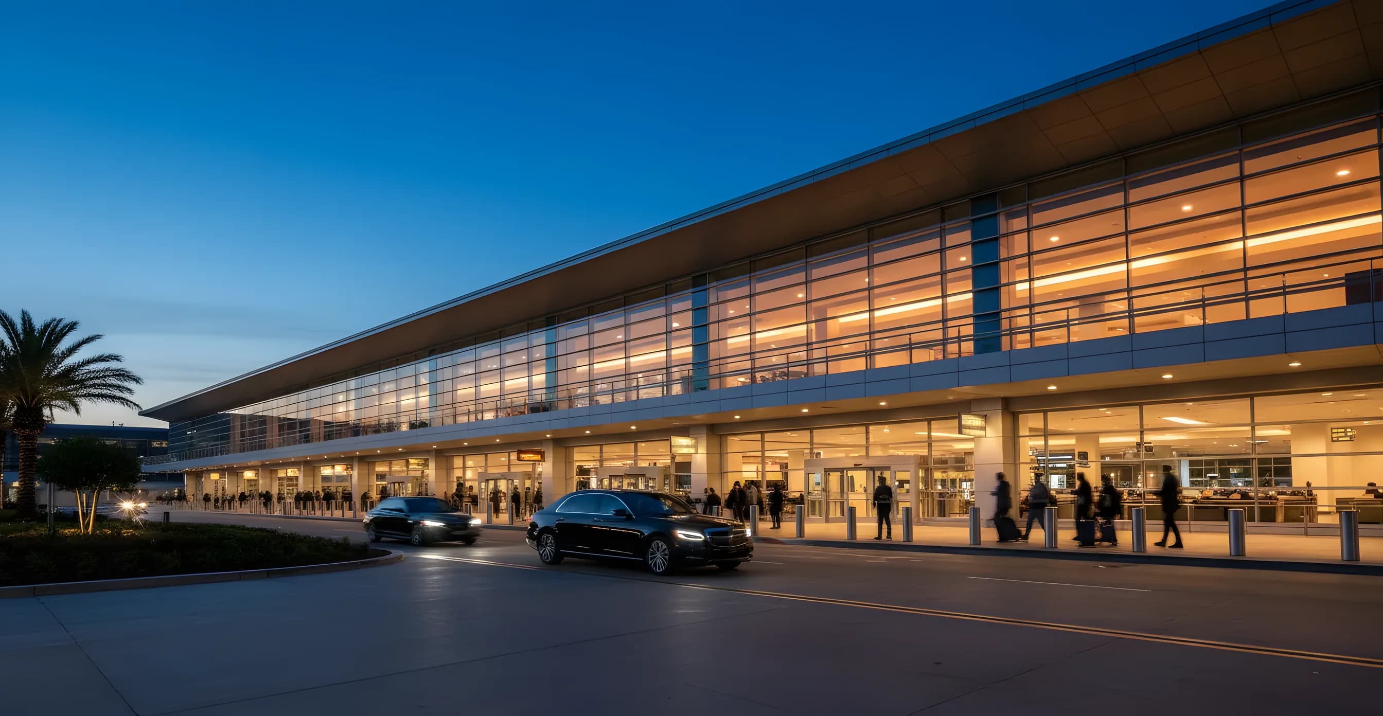 A modern glass airport terminal at dusk, with warm lights glowing from within and a car at the curb.