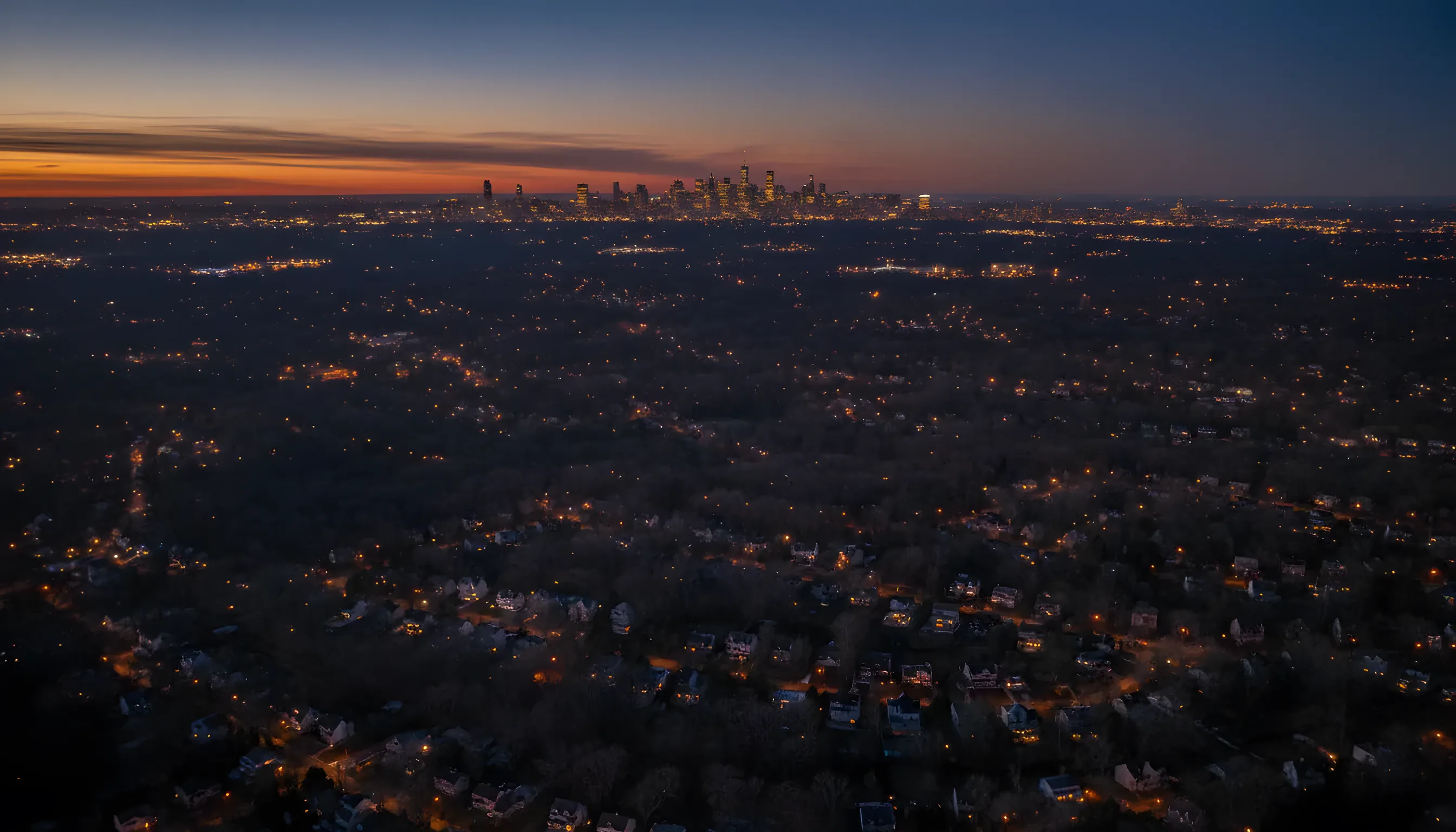 Aerial view from a helicopter flying over a suburban area towards a brightly lit city skyline at dusk.