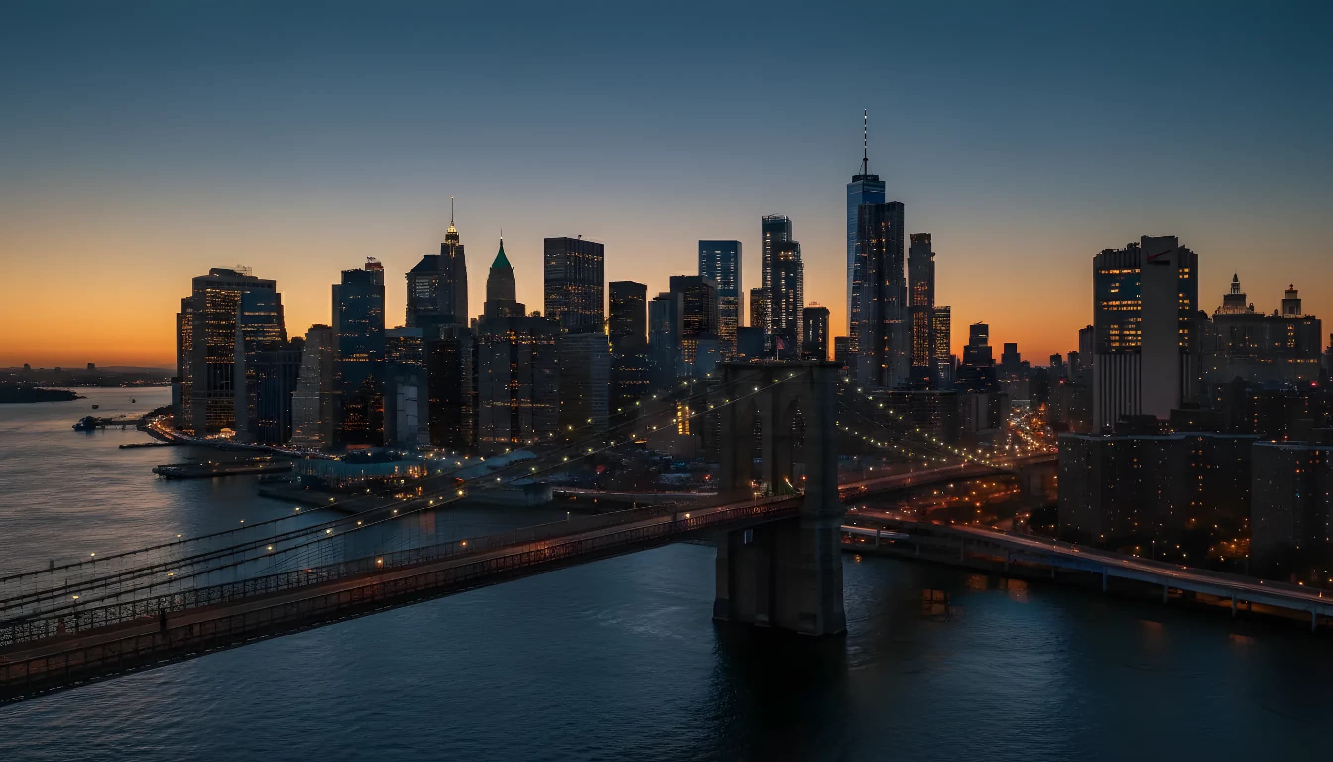 Aerial view of the Brooklyn Bridge and the Lower Manhattan skyline at dusk, with city lights beginning to glow.