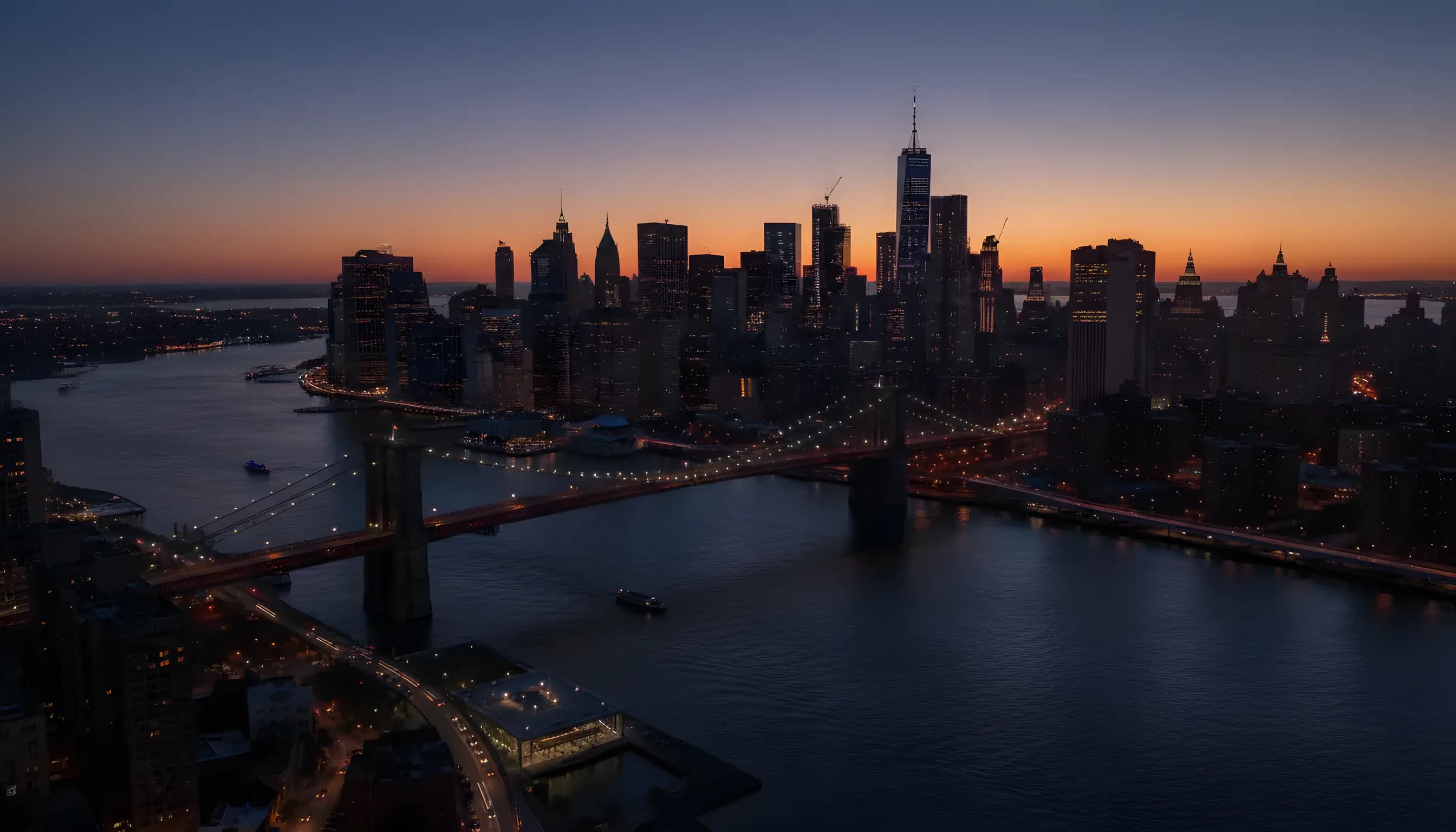 Sweeping aerial view of the lower Manhattan skyline at dusk, with city lights beginning to glow against a dark blue sky.
