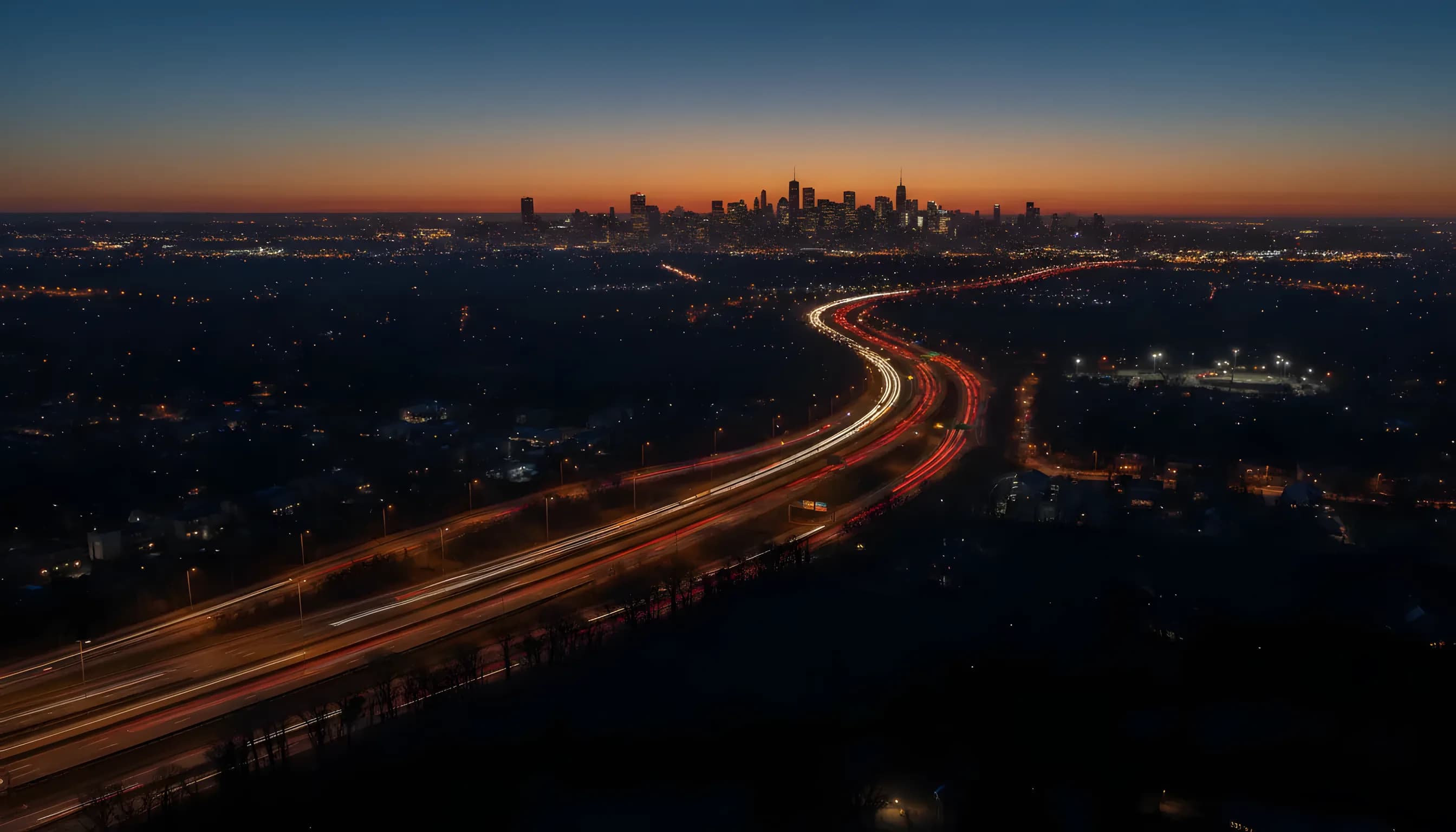 Aerial view of the distant Manhattan skyline at dusk, with light trails from highway traffic leading towards the city.